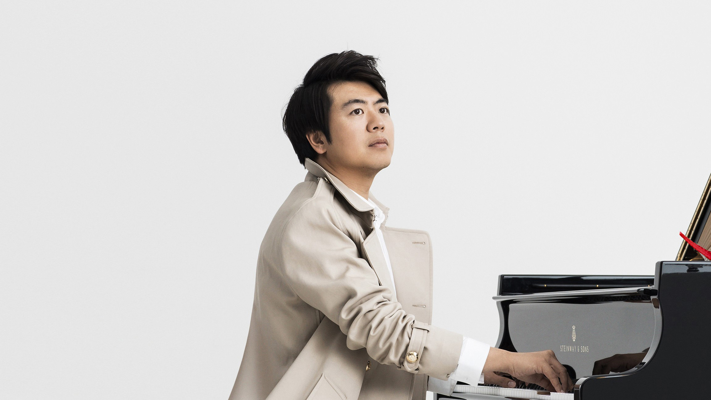 An Asian man, looking out, wearing a  white coat, playing the Deutsche Grammophon piano, a dark coloured piano with an accordion on the top.